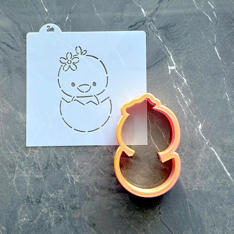 Puede incluir: Una plantilla de pl&aacute;stico blanco con un dise&ntilde;o de pollito con una flor en la cabeza y un cortador de galletas naranja a juego. El pollito est&aacute; saliendo de un huevo roto. La plantilla tiene una peque&ntilde;a pesta&ntilde;a con el n&uacute;mero 2.