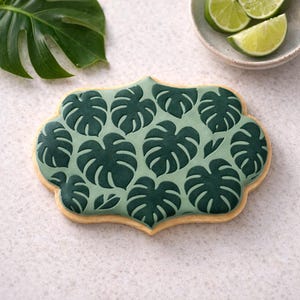 May include: A decorated cookie with a light green background and a pattern of dark green monstera leaves. The cookie has a light brown, scalloped edge. Sliced limes in a small bowl are in the background, along with a monstera leaf.