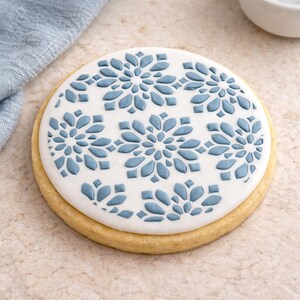May include: A round sugar cookie with white icing decorated with a blue snowflake pattern. The cookie has a golden-brown edge and is set against a neutral background.