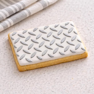 May include: A rectangular cookie with a white icing top decorated with a silver diamond plate pattern. The cookie has a golden-brown edge and sits on a light-colored surface. A gray and white striped towel is in the background.