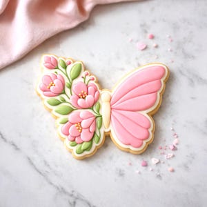 May include: A butterfly-shaped biscuit decorated with pink icing and floral details. The biscuit features a light pink wing design and a cluster of pink flowers with green leaves on the other wing. The biscuit is outlined in white icing and sits on a marble surface.