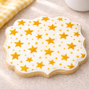 Puede incluir: Una galleta en forma de estrella con glaseado blanco y un patrón de estrellas y puntos amarillos. La galleta tiene un borde festoneado y una base marrón claro. Un dulce.