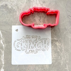 May include: A red plastic cookie cutter in the shape of a cloud sits above a white stencil. The stencil reads "in my GRINCH era" in a whimsical font. A small heart-shaped hole is cut into the stencil.