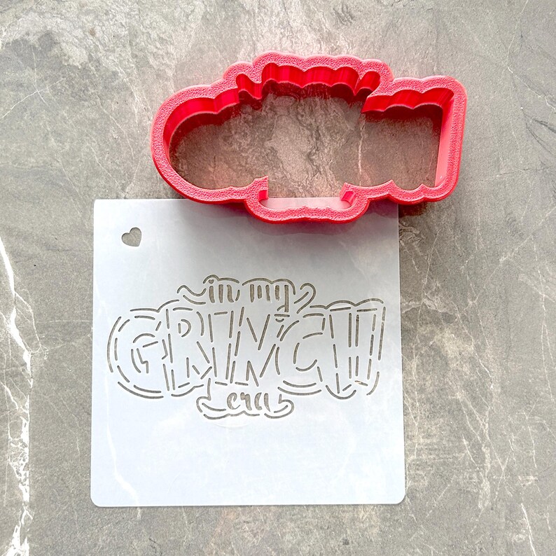 May include: A red plastic cookie cutter in the shape of a cloud sits above a white stencil. The stencil reads "in my GRINCH era" in a whimsical font. A small heart-shaped hole is cut into the stencil.