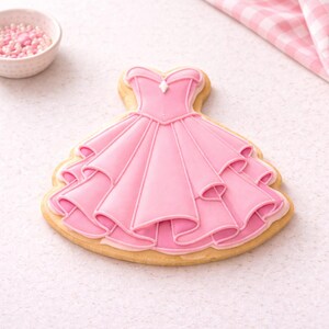 May include: A pink dress-shaped sugar cookie with white icing details. The cookie has a layered skirt design and a bodice with a small gem. A small bowl of pink sprinkles and a pink and white checkered cloth are in the background.