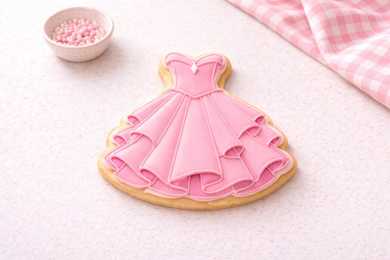 May include: A pink dress-shaped sugar cookie with white icing details. The cookie has a layered skirt design and a bodice with a small gem. A small bowl of pink sprinkles and a pink and white checkered cloth are in the background.