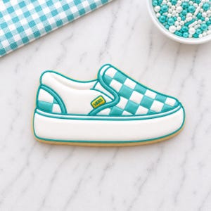 May include: A shoe-shaped cookie decorated to resemble a Vans sneaker. The cookie features a white sole, a teal and white checkered pattern, and a small yellow logo. The cookie is outlined in teal and brown.
