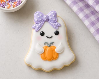 May include: A ghost-shaped sugar cookie decorated with white icing, featuring a light purple polka-dot bow on its head, black eyes, a small smile, and pink cheeks. The ghost is holding a small orange pumpkin with a grey stem in its center.