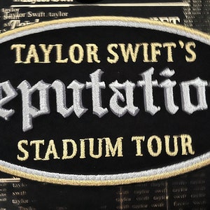 Può includere: Una toppa ovale nera con ricami bianchi e dorati. Il testo "TAYLOR SWIFT'S reputation STADIUM TOUR" è ricamato sulla toppa.