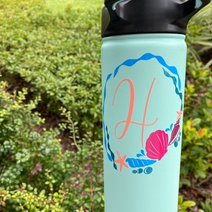 Puede incluir: Una botella de agua azul claro con un diseño de conchas blancas y azules y la letra "H" en color melocotón.