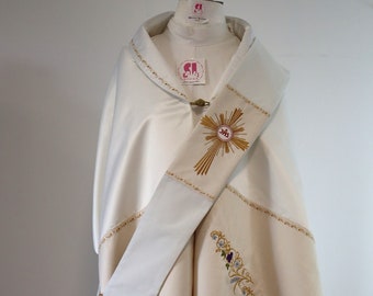 May include: A white liturgical vestment with gold embroidery and a gold cross with a circular center. The vestment has a white sash with gold trim and floral embroidery.