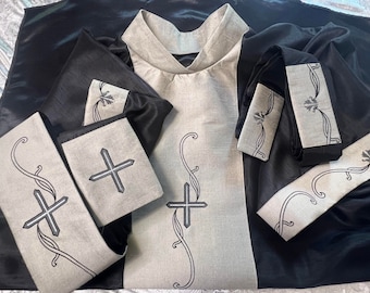 May include: A black and beige religious vestment set. The set includes a robe, stole, and cuffs, all featuring embroidered crosses and decorative scrollwork. The fabric has a subtle sheen, and the design is symmetrical and formal.