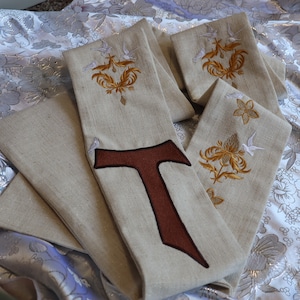 May include: A set of beige fabric stoles with embroidered designs. The stoles feature a brown cross, a floral design, and a bird design.