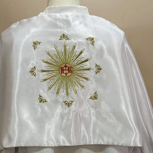 Könnte beinhalten: Weißes religiöses Gewand mit goldener Stickerei. Das Design zeigt einen Sonnenstrahl mit den Buchstaben IHS in der Mitte, umgeben von dekorativen Verzierungen. Der Stoff hat einen Satin-Glanz.