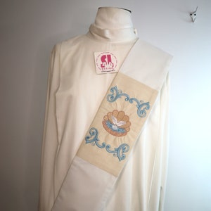 May include: White liturgical stole with a cream colored embroidered panel featuring a dove in a seashell with blue and gold accents. The stole has a label that reads "SI DESIGN".