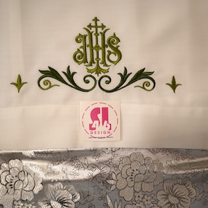 May include: White fabric with green embroidered floral design featuring the letters IHS with a cross above. A sticker with the text "S1 416 DESIGN" is placed on the fabric.