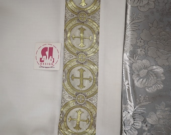 May include: White fabric with a gold and white trim featuring a repeating pattern of crosses and circles. The trim is on the right side of the fabric.