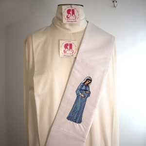 May include: White liturgical stole with an embroidered image of the Virgin Mary in blue and white. The stole has two pink tags with the text "SL DESIGN" on them.