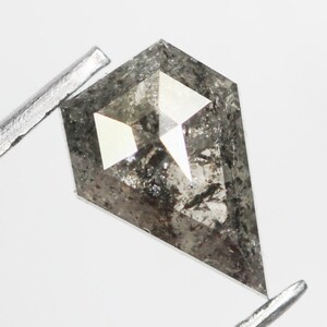 Può includere: Una pietra preziosa grigia, sfaccettata, a forma di diamante con una punta appuntita. La pietra ha una superficie ruvida e testurizzata ed è incastonata in una montatura argentata.