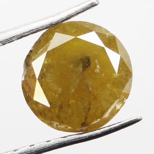 Op de afbeelding: Een ronde, gefacetteerde, geelbruine diamant, vastgehouden door een pincet. De edelsteen heeft een briljantslijpsel met meerdere facetten die licht reflecteren. De kleur van de diamant varieert van licht- tot donkergeelbruin, met zichtbare insluitsels.