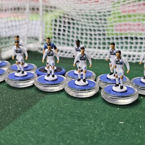 May include: Miniature football players in white kits with black details stand on blue bases. A white goal net and posts are visible in the background, indicating a tabletop football game.