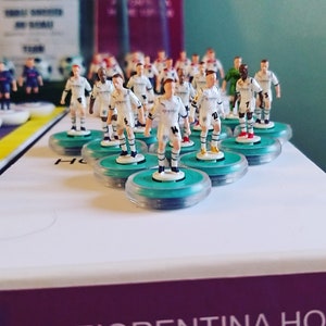 May include: A collection of miniature soccer player figurines in white jerseys with light blue accents, standing on teal and clear circular bases. The figures are arranged on a white surface, suggesting a board game or collectible item.