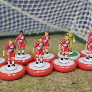 May include: A collection of miniature football player figurines standing on circular red and white bases. Each figure wears a red and white jersey with the word GRILLA printed on the front. The players are arranged in front of a white mesh goal net.