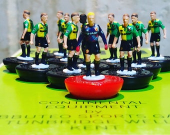 Könnte beinhalten: Eine Ansammlung von Miniatur-Fußballspielerfiguren in grünen, gelben, schwarzen und roten Trikots. Die Spieler stehen auf schwarzen Sockeln, mit einem roten Sockel im Vordergrund. Das Bild hat einen hellgrünen Hintergrund mit Text.