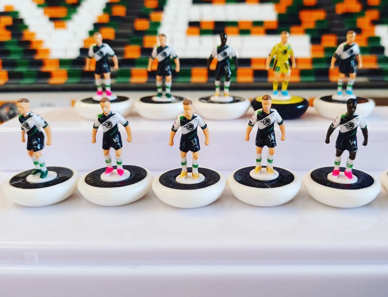 May include: A collection of miniature soccer player figurines. The players are dressed in white, black, and green uniforms, standing on white bases with black and white details. Some players have different skin tones and colored shoes.