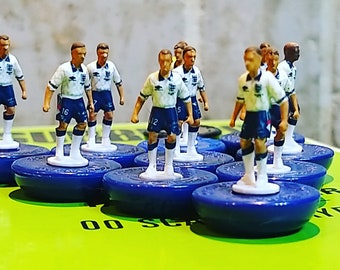 Könnte beinhalten: Miniaturfußballspieler-Figuren in weißen und blauen Trikots, montiert auf blauen Sockeln. Die Figuren tragen die Uniform der englischen Mannschaft. Sie sind auf einer grünen Oberfläche angeordnet, vermutlich einem Spielbrett.