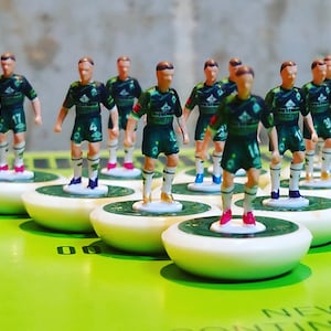 Puede incluir: Figuras en miniatura de jugadores de fútbol con uniformes verdes y blancos. Las figuras están sobre bases blancas con círculos verdes. Están dispuestas sobre una superficie verde, sugiriendo un juego o exhibición.