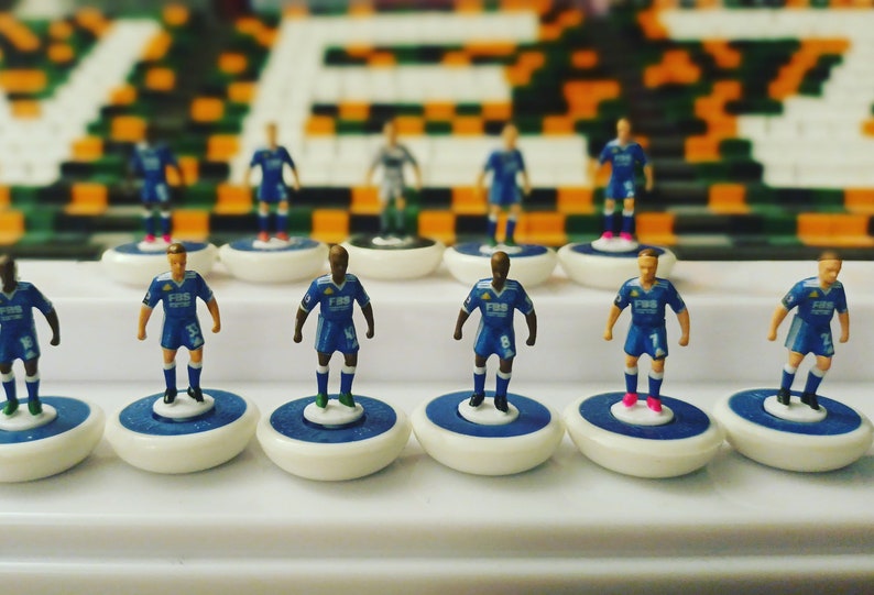 May include: A collection of miniature soccer player figurines in blue uniforms with white and blue accents. The players stand on white and blue bases, arranged in rows. The background is a blurred image of a stadium.