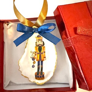 Pode incluir: Enfeite de Natal de concha de ostra pintado à mão com um soldado quebra-nozes em ouro, azul e preto. O enfeite tem uma fita dourada e um laço azul, apresentado numa caixa de presente vermelha.