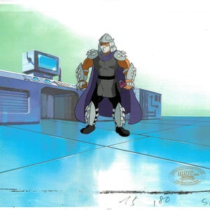 May include: Animation cel of the Shredder from Teenage Mutant Ninja Turtles. The character is in a gray and purple outfit with a helmet and cape. He is standing on a blue surface with a control panel and a computer monitor in the background.