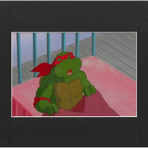 May include: A vintage animation cel featuring a green anthropomorphic turtle character with a red eye mask, wearing a brown chest plate, and resting in a bed with a pink blanket. The character has its eyes closed and mouth slightly open.