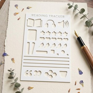 May include: A white rectangular plastic stencil labeled READING TRACKER with various cutouts including an open book, headphones, speaker, stars, hearts, leaves, geometric shapes, horizontal lines, quotation marks, and a honeycomb pattern.