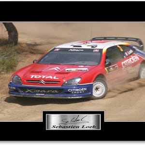 May include: A red, white, and blue Citroën Xsara rally car speeds across a dirt track, kicking up dust. The car features sponsor logos, including "TOTAL" and "CITROËN SPORT." A framed image with the signature of Sébastien Loeb is at the bottom.