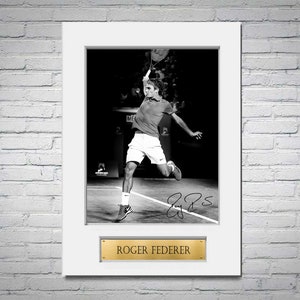 Può includere: Fotografia in bianco e nero incorniciata di Roger Federer in azione durante una partita di tennis. L'immagine è firmata e include una placca dorata con il nome "Roger Federer". La fotografia è montata su un passe-partout bianco.