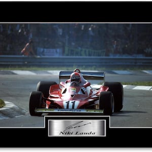 May include: A framed photograph of a red Formula 1 racing car, number 11, driven by Niki Lauda. The car is on a racetrack, with a crowd in the background. A silver plaque with "Niki Lauda" is at the bottom of the image.