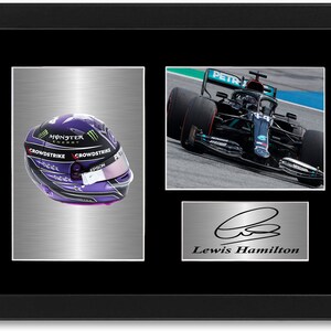 May include: Framed artwork with a Formula 1 racing helmet, a race car, and a signature. The helmet is purple and black, with sponsor logos. The signature reads "Lewis Hamilton". The car is in motion.