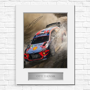 May include: Framed photograph of a rally car in action, splashing through water. The car is blue, red, and white, with sponsor logos. The frame is white, with a silver nameplate that reads "OTT TANAK" and an autograph.