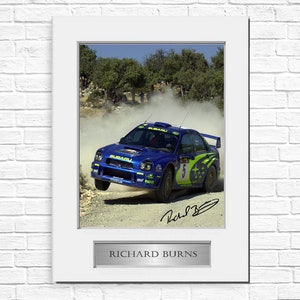Puede incluir: Fotografía enmarcada de un coche de rally Subaru azul en acción, levantando polvo en un camino de tierra. El coche tiene detalles verdes y amarillos y el número 5. Incluye una firma y una placa que dice "RICHARD BURNS".