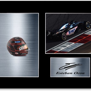 Może przedstawiać: Oprawiony wydruk przedstawiający kask wyścigowy Formuły 1 z czerwonymi akcentami i samochód wyścigowy na torze. Wydruk zawiera również podpis i nazwisko "Esteban Ocon". Kask ma logo "BWT".