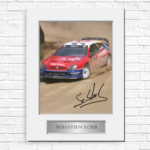 May include: Framed photograph of a red, white, and blue Citroen Xsara rally car in action, kicking up dust. The image includes the signature of Sebastien Loeb and a plaque that reads "SEBASTIEN LOEB". The photograph is set in a white frame.
