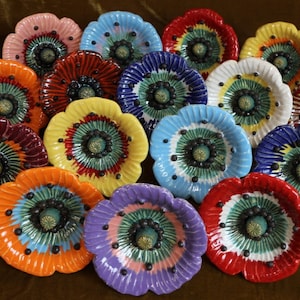 Könnte beinhalten: Eine Sammlung von 15 Keramik-Mohnblumen-Dekorationen in verschiedenen Farben, darunter Rot, Blau, Gelb, Orange und Lila. Jede Mohnblume hat eine schwarze Mitte mit einem grünen und weißen Design.