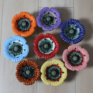 Könnte beinhalten: Acht Keramik-Mohnblumen-Schalen in verschiedenen Farben, darunter Rot, Blau, Gelb, Orange und Lila. Jede Schale hat eine grüne Mitte mit schwarzen Punkten und eine erhabene Mitte.