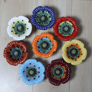 Könnte beinhalten: Acht Keramik-Mohnblumen-Schalen in verschiedenen Farben, darunter Rot, Blau, Gelb, Orange und Weiß. Jede Schale hat ein zentrales schwarzes und grünes Design mit einer erhabenen Mitte.