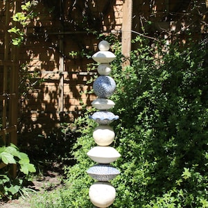 Könnte beinhalten: Eine hohe Gartenskulptur aus gestapelten weißen, blauen und gemusterten Keramikkugeln und -scheiben. Die Skulptur ist auf einer Metallstange montiert und vor dem Hintergrund von üppigem Grün und einer Backsteinmauer platziert.