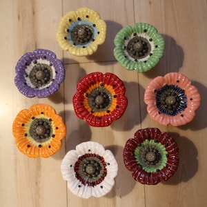 Könnte beinhalten: Satz von neun Keramik-Schubladengriffen in Form von Mohnblumen in verschiedenen Farben, darunter Rot, Gelb, Grün, Lila, Orange, Rosa, Weiß und Burgunder. Jeder Griff hat eine schwarz-weiße Mitte mit einem braunen Knopf.