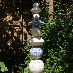 Könnte beinhalten: Eine hohe Gartenskulptur aus gestapelten Keramikkugeln und -scheiben. Die Skulptur weist eine Mischung aus weißen, blauen und gemusterten Elementen auf, mit einer rissigen Textur auf einigen der Kugeln. Sie steht vor einem Hintergrund aus grünem Laub und einer Backsteinmauer.
